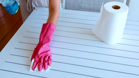 a person wearing pink rubber gloves cleans a white slatted table using cleaning spray and a paper towel in a brightly lit indoor setting