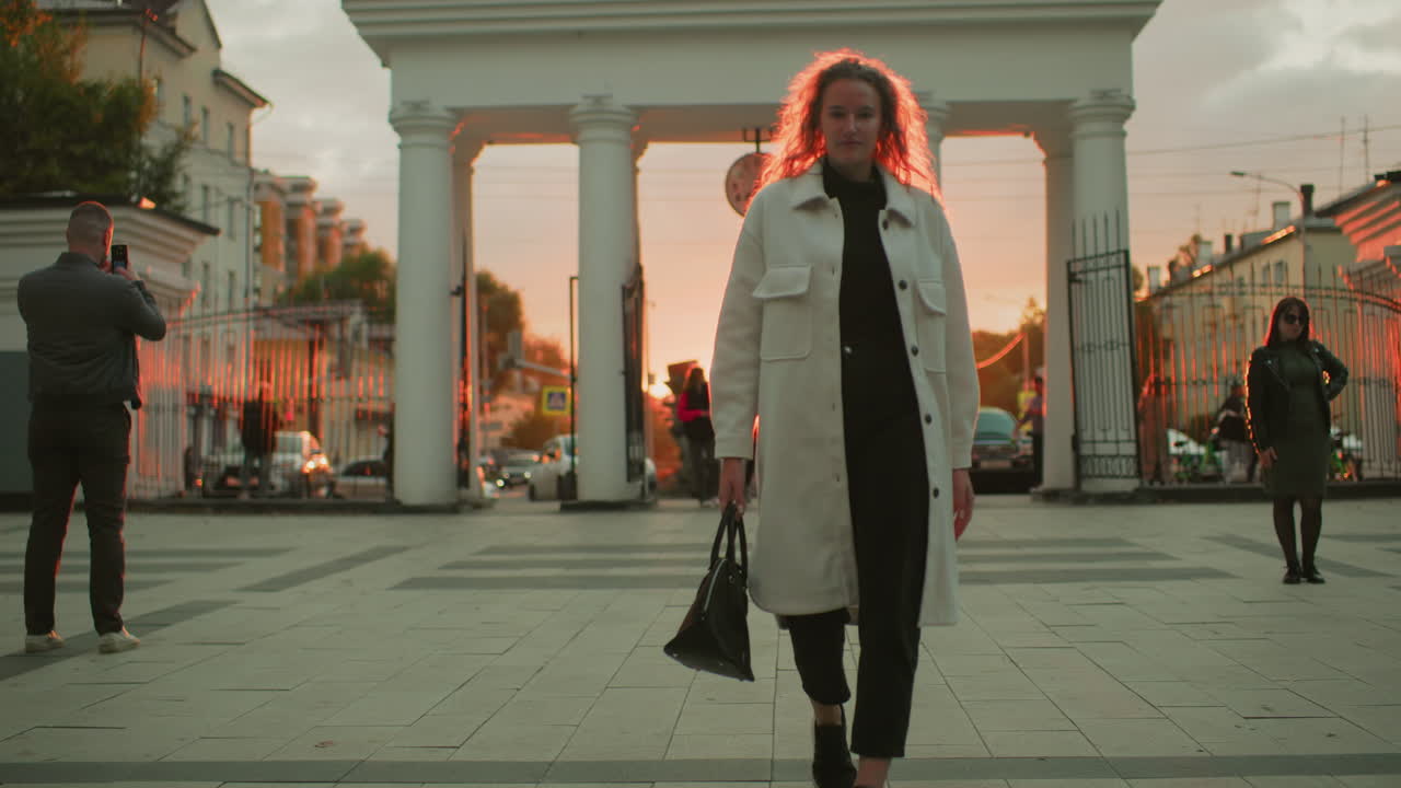 business woman in white coat carrying handbag walking confidently under urban archway during sunset with warm red sunlight shining from behind in busy city setting with people and cars in background