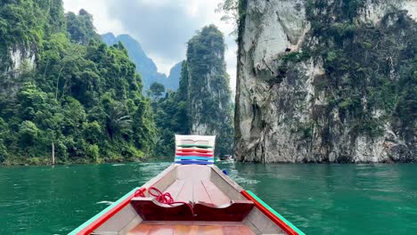 navegando por el lago khao sok en un barco de cola larga, el espectador observa la parte delantera del barco con tela de colores, aguas verdosas y montañas cubiertas de vegetación acercándose a las rocas de khao sam kloe