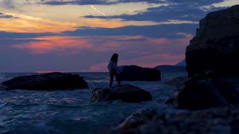 Chica-Caminando-Por-Acantilados-Sobre-Agua-De-Mar-Con-Un-Hermoso-Fondo-De-Colorido-Atardecer-En-El-Cielo-Nublado