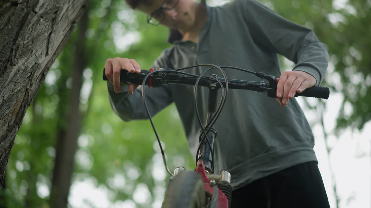 niño con gafas sostiene el manillar de una bicicleta estacionada en un campo cubierto de hierba, agarra el manillar con firmeza, comprobando la funcionalidad de los frenos mientras está de pie, la bicicleta está estacionada cerca de un árbol
