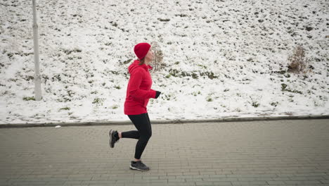 side view of coach jogging outdoors on interlocked pavement next to snowy hill, winter scene features athletic motion, vibrant red hoodie, serene environment, and natural snowy hillside