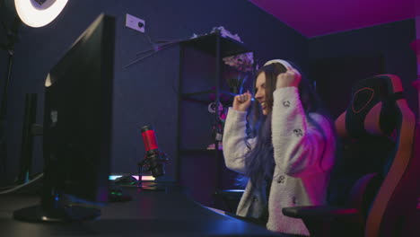 young female gamer in cozy sweater adjusts headphones while preparing for interactive gameplay session, seated at modern desk with microphone, monitor, and gaming chair under moody ambient lighting