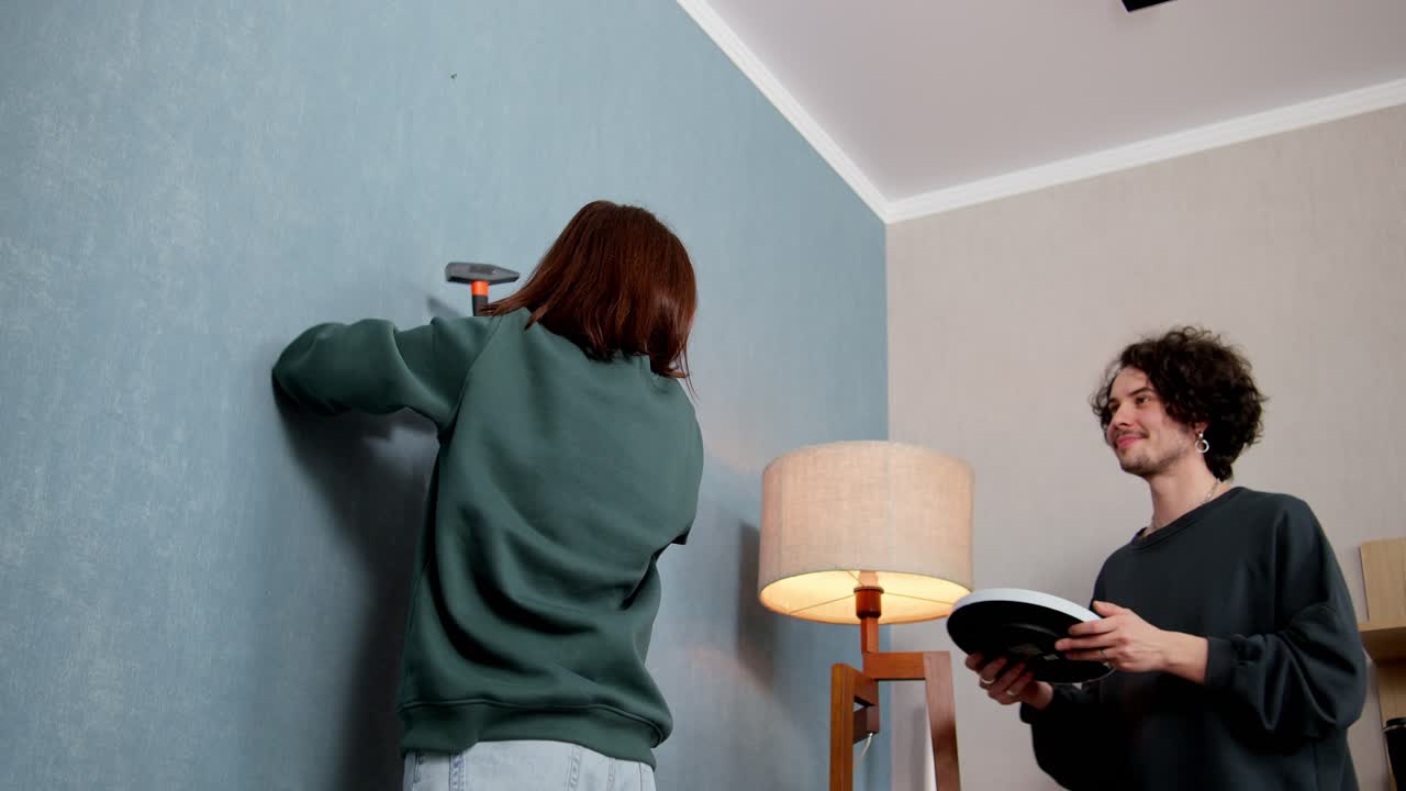 초록색 재을 입은 갈색 소녀가 파란 벽에 못을 고 현대적인 아파트에서 남자친구의 지원을 받아 시계를 매달고 있습니다.