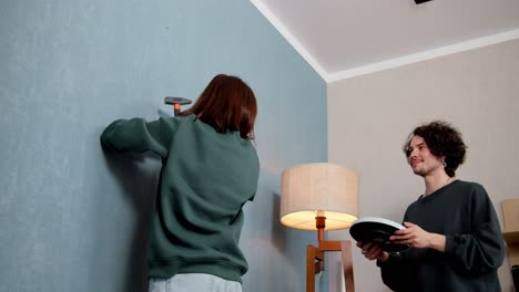 A-brunette-girl-in-a-green-jacket-hammers-a-nail-into-a-blue-wall-and-hangs-a-clock-with-the-support-of-her-boyfriend-at-home-in-a-modern-apartment