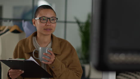 a woman wearing glasses and a brown jacket is standing in an office and holding a clipboard and a mug. she is looking at a screen and seems to be in a meeting or presentation.