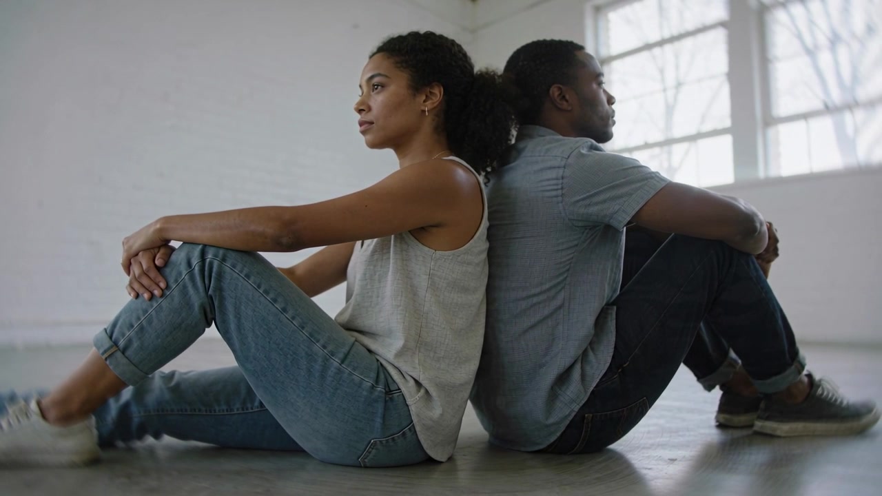 Settling back to back, woman left, man right adjusting for comfort wearing jeans, sneakers, window