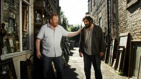 in a narrow alley lined with vintage items and framed artwork, two men engage in a conversation, highlighting their contrasting personalities and surroundings