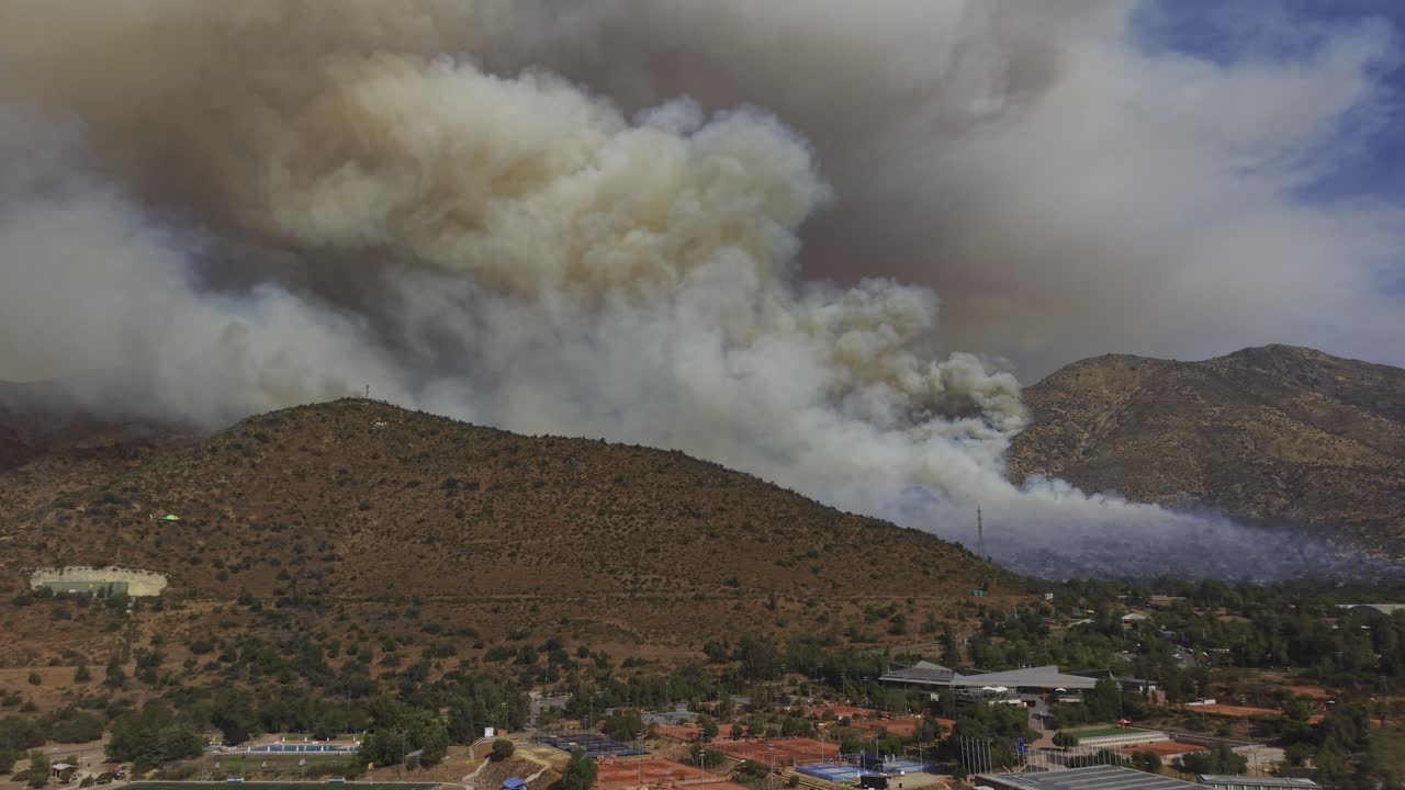 Vista aerea cinematografica con drone di un enorme incendio che produce spesse colonne di fumo sul pendio di una montagna, direttamente dietro un quartiere residenziale a Santiago, Cile, 4K