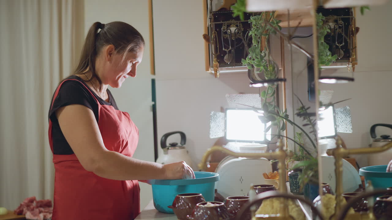 Mulher de avental vermelho mexendo tigela na pia, com luz de fundo brilhante e plantas penduradas, sorrindo enquanto mede e mistura, cozinheira preparando refeição em cozinha aconchegante, momento de vida casual