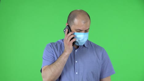 portrait of a middle aged men in medical mask talking on his smartphone. balding male in blue shirt posing on green screen in the studio. close up