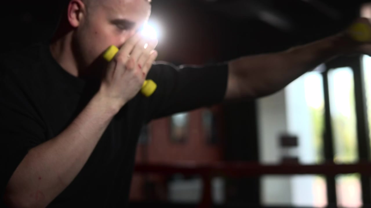 Solitary boxer stands inside dim ring with raised hands, yellow wraps near face, red ropes framing scene, strong side light shaping focused training mood inside industrial gym interior