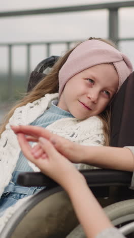 caring woman strokes hand of little daughter with leg injury on walk. girl covered with knitted shawl smiles looking at mother sitting in wheelchair after surgery