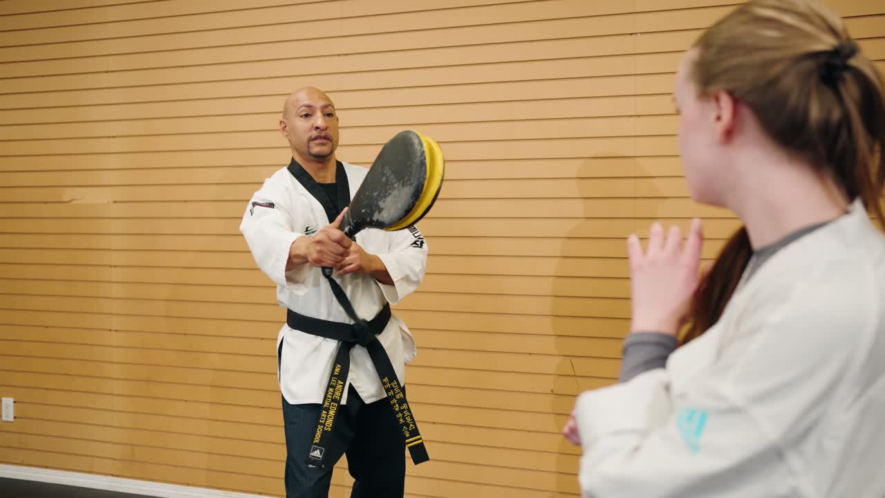 A Male Martial Arts Karate Master Holds a Kick Target for a Student Against a Wooden Board Background Inside a Professional Combat Training Gym