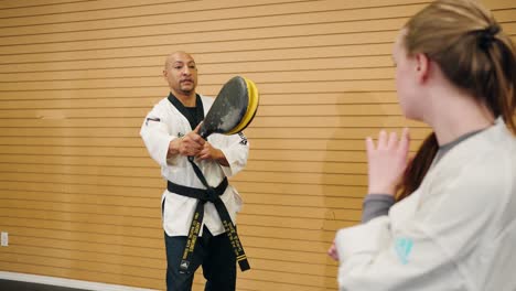 a male martial arts karate master holds a kick target for a student against a wooden board background inside a professional combat training gym