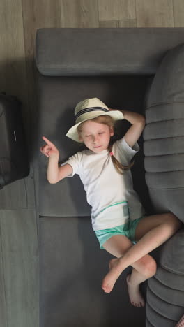 toddler walks past elder sister resting on sofa and big tourist suitcase. children spend time in hotel room after arrival upper view