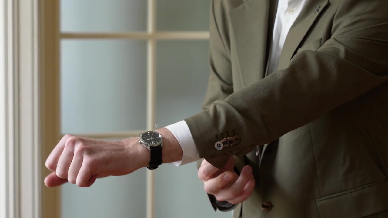 Close-up shot of a man in a stylish olive suit adjusting his wristwatch in a softly lit, elegant room with windowed doors. Focus highlights confidence, business style, and refined details.