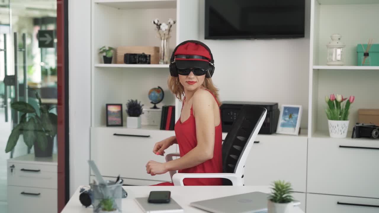 la mujer con un vestido rojo y gafas de sol se sienta en una mesa en una oficina luminosa, escuchando música en auriculares inalámbricos, bailando en una silla