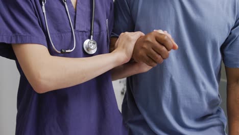 midsection of diverse female doctor supporting male patient walking in hospital