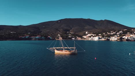 a stunning aerial view of a drone flying over a wooden sailboat anchored in a beautiful bay, with a charming village in the background