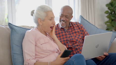 una feliz pareja biracial sentada en el sofá y usando una computadora portátil en casa, en cámara lenta