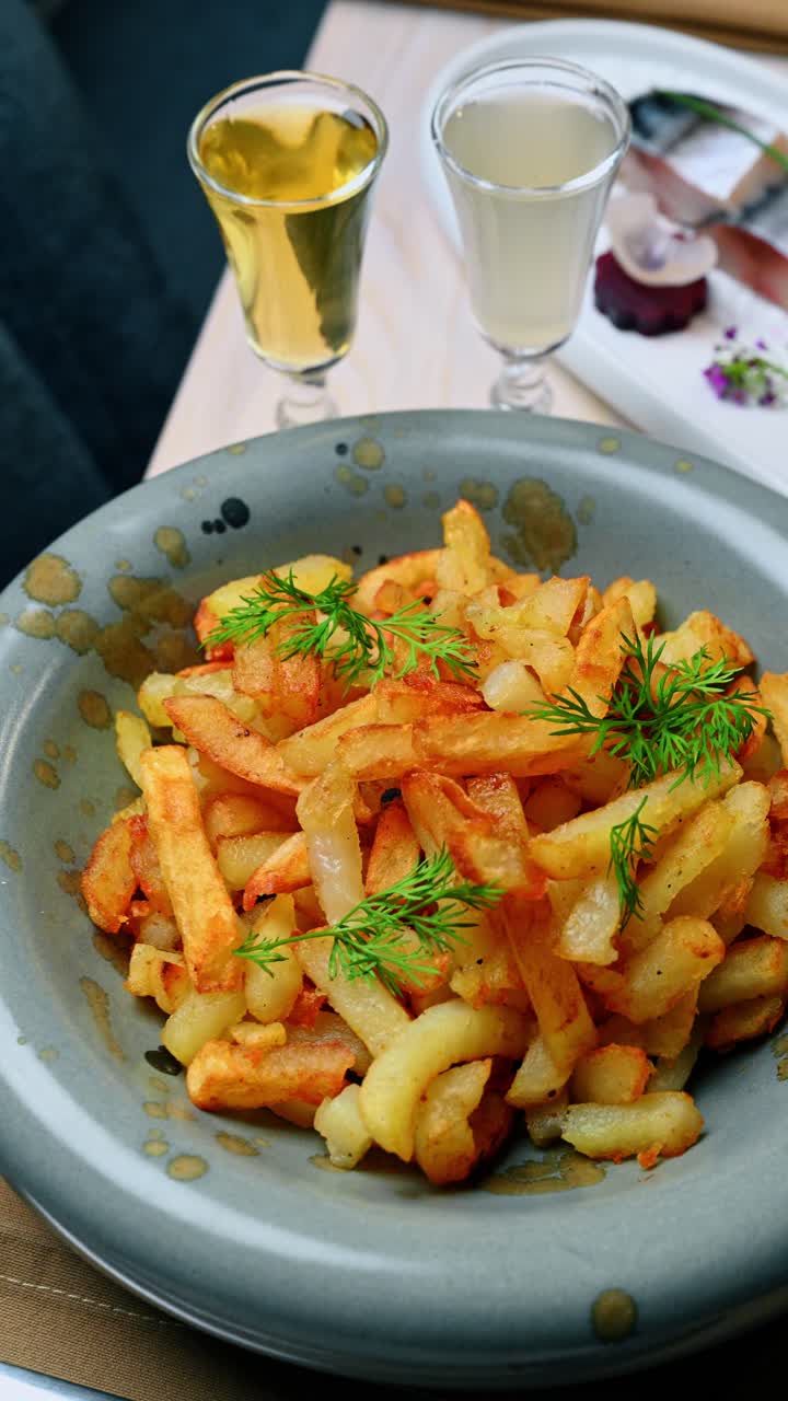 Fried potatoes with dill served in bowl with drinks. Top view of golden fried potatoes garnished with fresh dill in a ceramic bowl next to two shot glasses