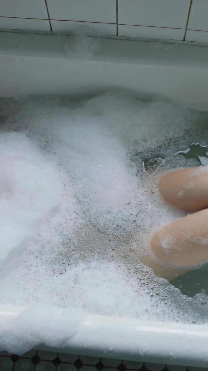 Video verticale: Gambe nude che si muovono spostando la schiuma nella vasca da bagno durante il bagno, rivelando lo scarico, con spazio per il testo.
