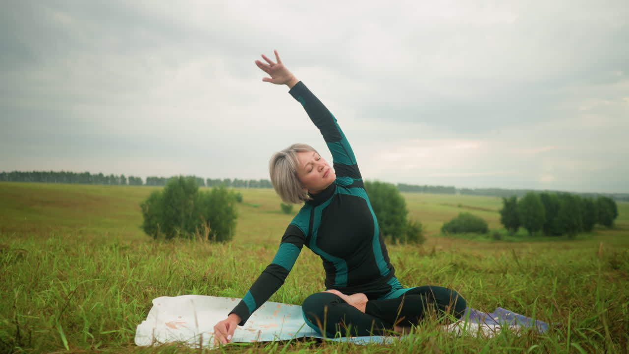 Woman with grey hair seated on yoga mat, practicing side bend pose with arms extended, eyes closed, and sitting in accomplished pose, under cloudy skies, with trees in the distance