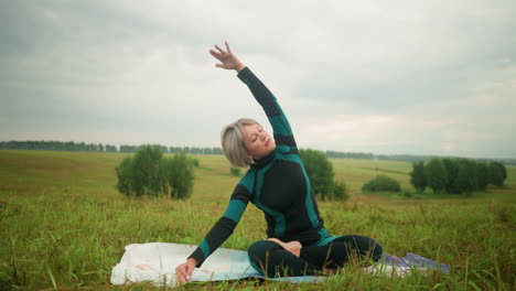 woman with grey hair seated on yoga mat, practicing side bend pose with arms extended, eyes closed, and sitting in accomplished pose, under cloudy skies, with trees in the distance