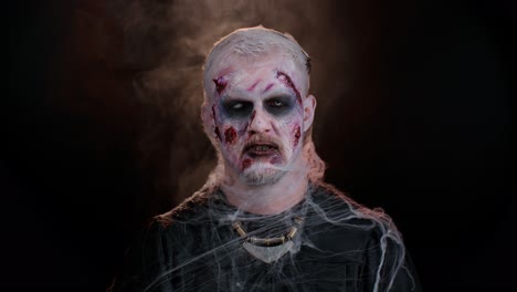 sinister devil man with scary halloween dead zombie makeup in costume making faces, trying to scare