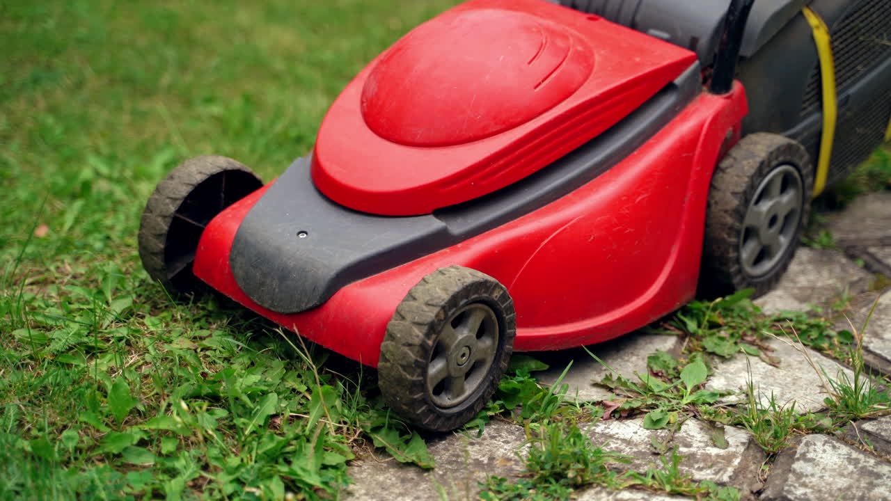 Mowing garden care work tool. Lawn mower cutting green grass near the tile track in the yard in summer. Red grass mower works. Close-up.