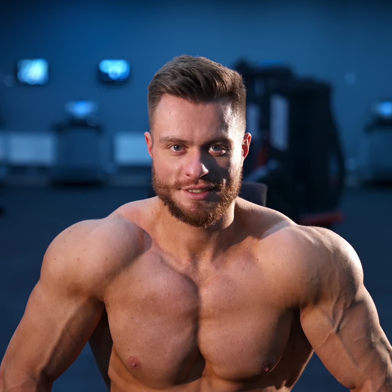 Male sportsman with naked torso and big muscles sits on training chair in modern gym. Male beauty concept, Slow motion