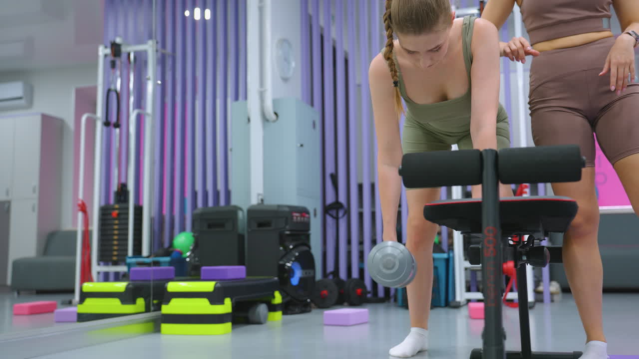 트레이너는 덤벨 행 연습을 수행하는 동안 자세를 안내하기 위해 클라이언트 등에 손을 놓습니다. 집중된 훈련생은 체조 장비와 활기찬 배경과 함께 결단력으로 무게를 들어 올립니다.