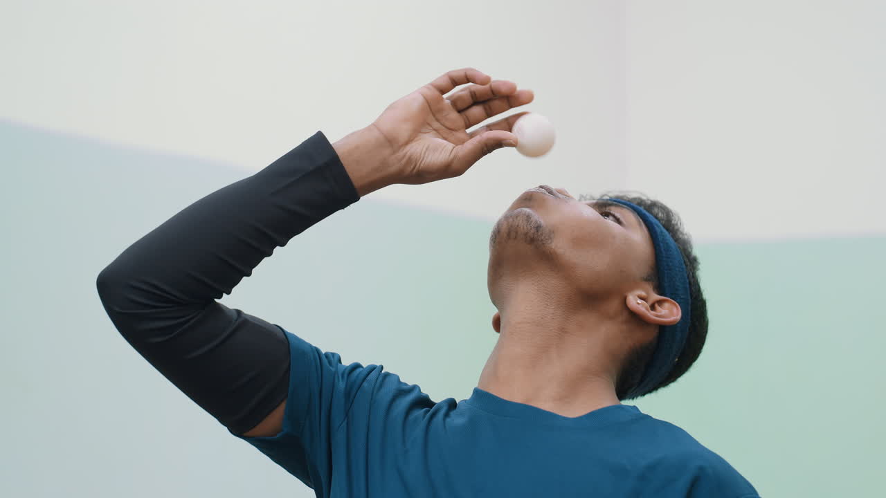 파란색 셔츠와 머리띠를 한 남자가 숨을 이용하여 테니스 공을 공중에 여러 번 균형을 잡으려고 집중하고 있어요. 장난기 넘치는 순간 동안 결연함과 끈기를 보여주는 집중된 표정이 인상적이네요.