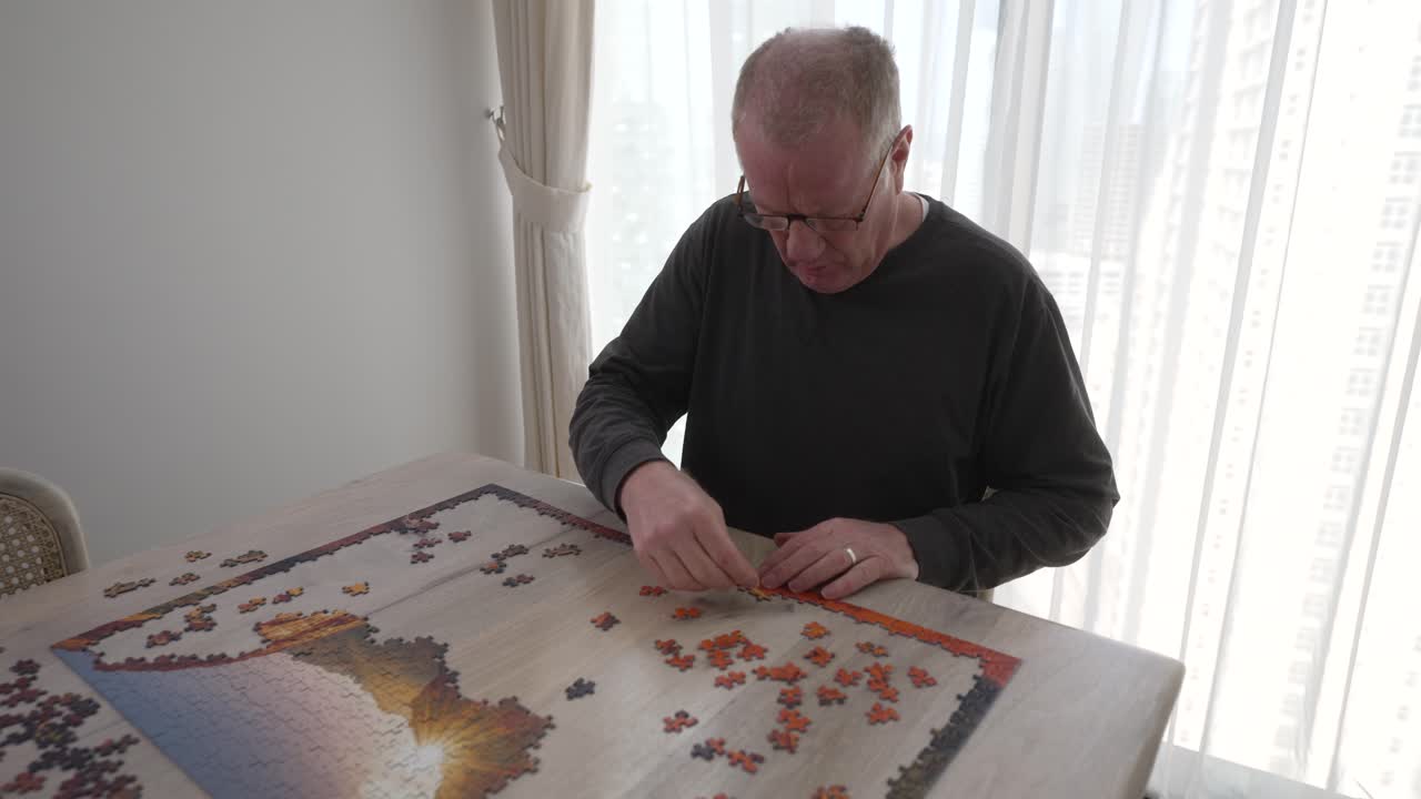Mature man with focused intent to put a large picture puzzle together in his lonely home