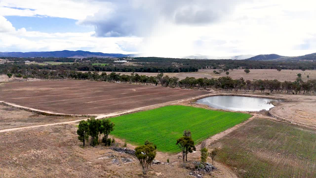 Drone footage glides over a rural landscape, beginning at a stone pyramid and moving across green fields, trees, and a farm dam under cloudy daylight