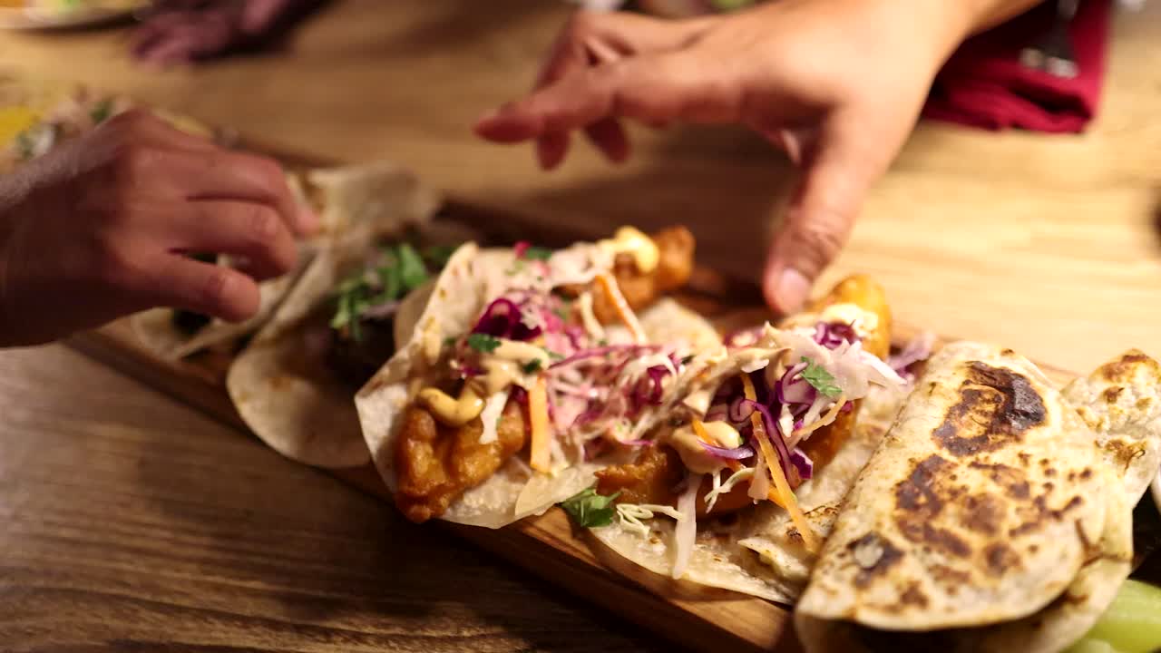 Multiple hands reach for assorted grilled soft shell tacos on a wooden board in a warmly lit restaurant setting, emphasizing shared dining and vibrant food presentation