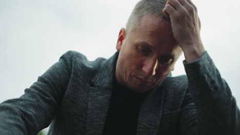 a close view of a man in a gray jacket and blue jeans sitting on a railway track, holding a handgun with his head bowed and hand resting on his head, with a blurred green forest