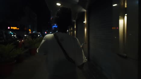 Handheld-dolly-shot-of-a-young-indian-woman-dressed-in-white-t-shirt-and-a-fanny-pack-while-walking-through-the-streets-at-night-looking-at-the-street-lights