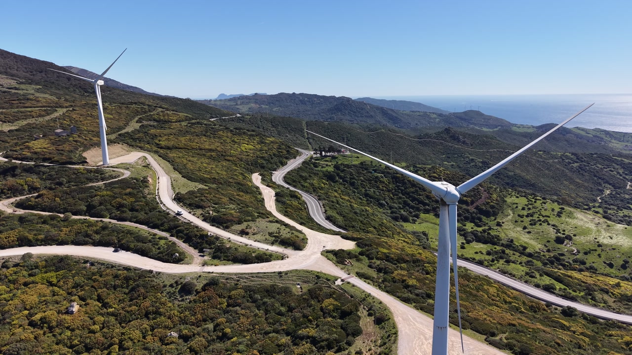 Aerial orbit of a wind turbine showing winding mountain roads and a dense wind farm network under a clear blue sky in southern Spain.