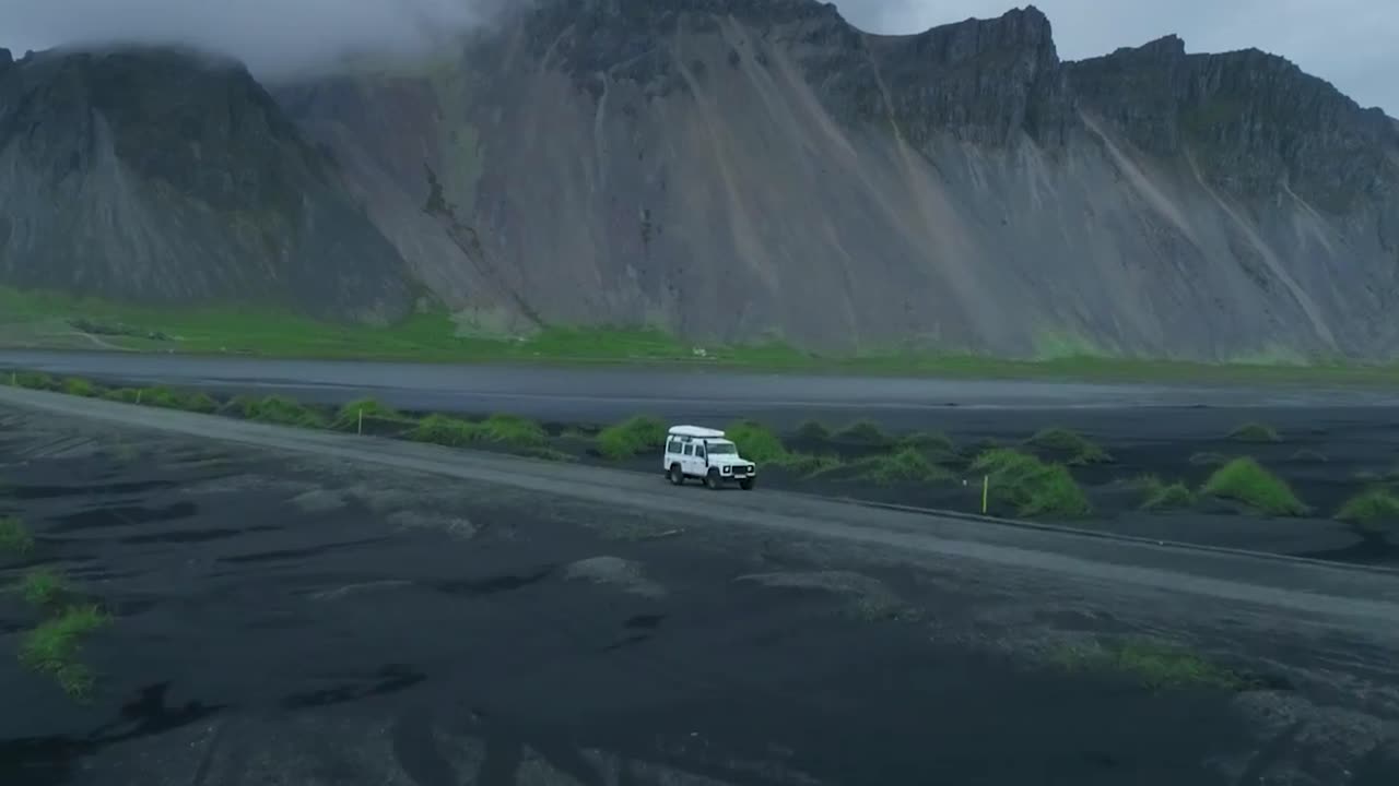 Aerial drone footage gliding towards and closer to a white 4x4 truck parked on a desolate and muddy road between big mountains at Greenland or Iceland during a cloudy and misty day. Moss and grass.