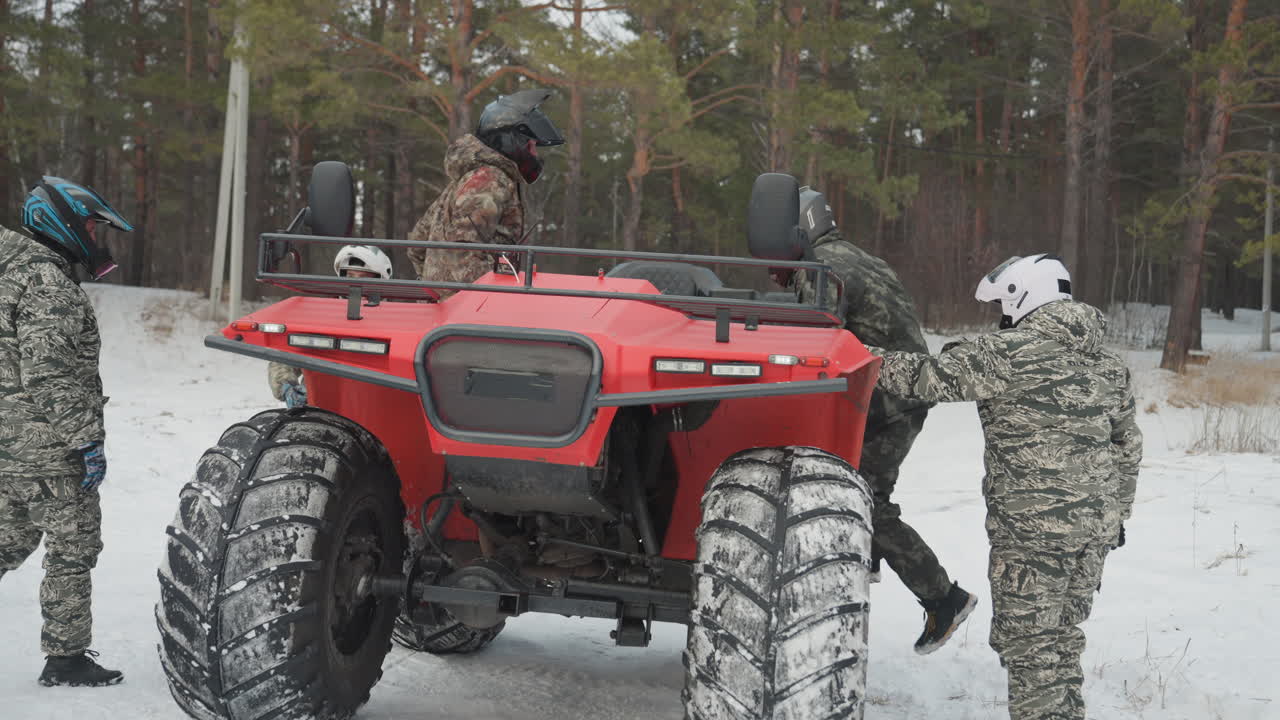 L'equipaggio ispeziona un quad rosso in una pineta innevata prima della partenza. Un meccanico controlla il motore, stringe i bulloni, la squadra sale sul veicolo e fissa l'attrezzatura, con i caschi indossati, le mani sulla chiave inglese, preparazione concentrata e robusta.