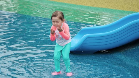 cute toddler girl in aquapark jumps in shallow puddle pool happily like a frog, splashes water in eyes and wipes face with hands playing at water park playground