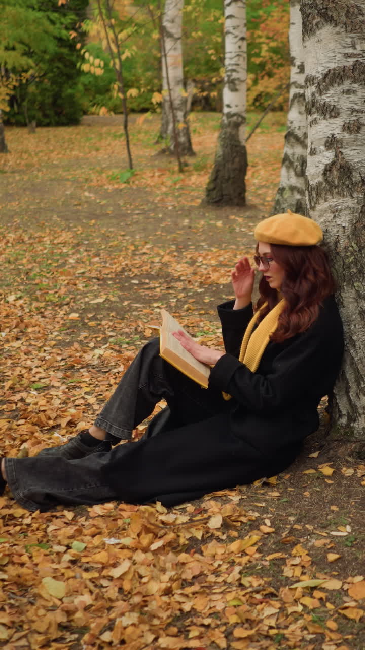 dama elegante se sienta en la soledad apoyándose en el árbol, ajustando las gafas mientras lee el libro pensativo, las hojas de otoño cubren el suelo, rodeando el entorno pacífico del bosque