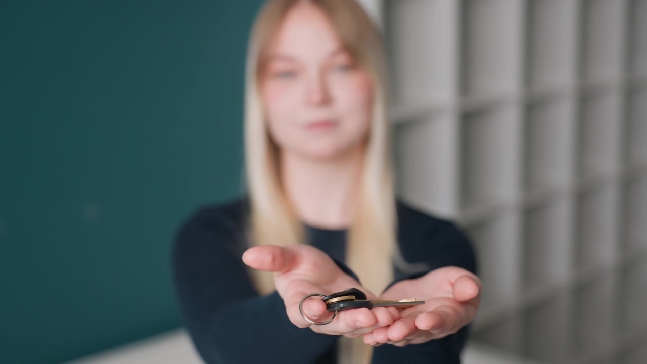Una donna bianca offre le chiavi a palmo aperto all'interno di un appartamento vuoto con parete verde acqua e scaffalature. L'inquilino accetta le chiavi della nuova casa con un sorriso gentile, in una scena a fuoco morbido che simboleggia il trasloco e l'inizio di un nuovo trasferimento.