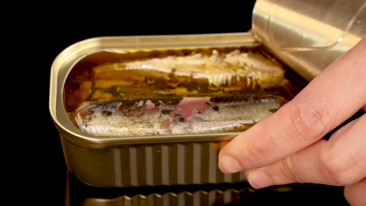 A close-up sequence shows a hand methodically peeling back the lid of a sardine can packed in oil under bright, even lighting with a static camera angle