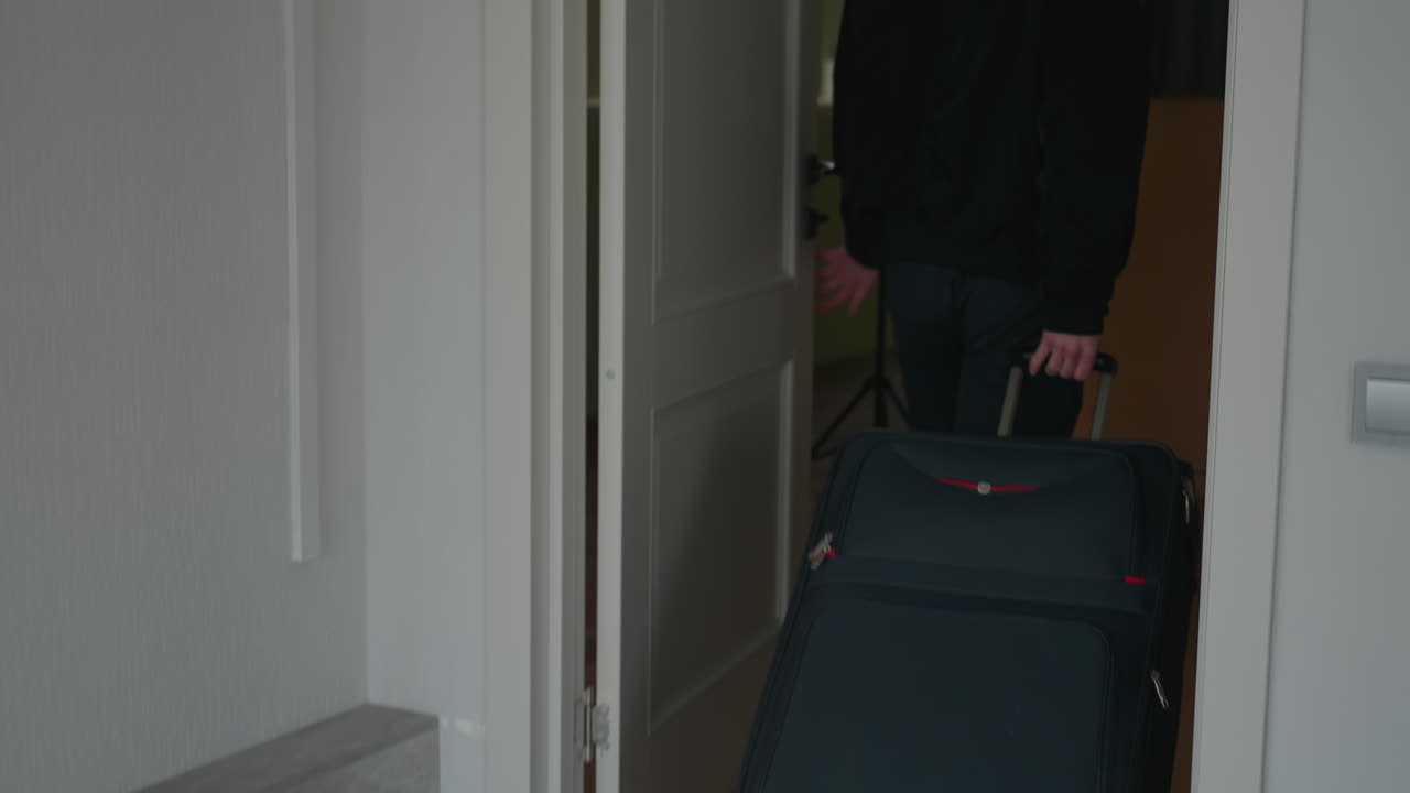 Rear view of man returning from journey rolling wheeled suitcase through doorway into room and closing door behind him with visible suitcase wheels on neutral toned floor