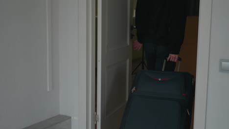 rear view of man returning from journey rolling wheeled suitcase through doorway into room and closing door behind him with visible suitcase wheels on neutral toned floor