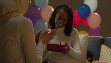 woman giving female friend birthday present at home with party balloons in background