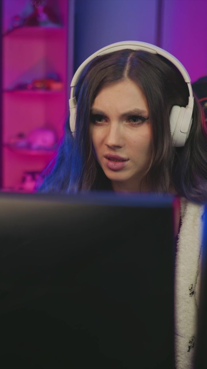 Una giovane donna con cuffie bianche è concentrata sullo schermo del computer davanti a sé. Lo sfondo presenta scaffali rosa con vari oggetti e l'illuminazione è prevalentemente viola e blu, creando